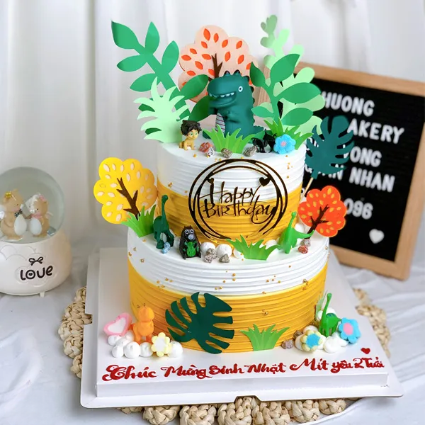 Bánh kem 2 tầng với khủng long đứng trên đỉnh và rừng cây phía dưới.
